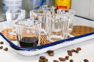 A white tray holding six empty shot glasses and one shot glass with a dark liquid, surrounded by scattered coffee beans.
