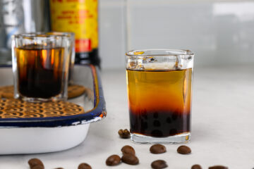 A shot glass with a layered coffee drink and tapioca pearls, surrounded by coffee beans on a white surface, with another glass and a bottle in the background.