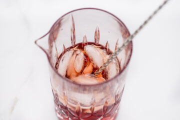A glass mixing vessel filled with a reddish liquid, ice cubes, and a metal stirring rod on a white surface.
