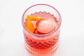 A glass of red cocktail with a large clear ice sphere and an orange peel garnish, set on a white background.