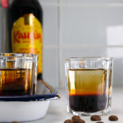 A glass of layered coffee liqueur cocktail sits on a counter with coffee beans, another glass, and a bottle of Kahl&uacute;a in the background.