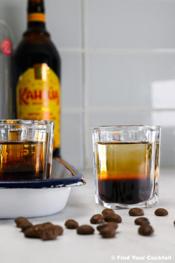 A glass of layered coffee liqueur cocktail sits on a counter with coffee beans, another glass, and a bottle of Kahl&uacute;a in the background.