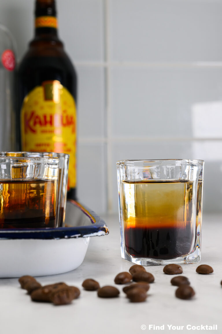 A glass of layered coffee liqueur cocktail sits on a counter with coffee beans, another glass, and a bottle of Kahlúa in the background.