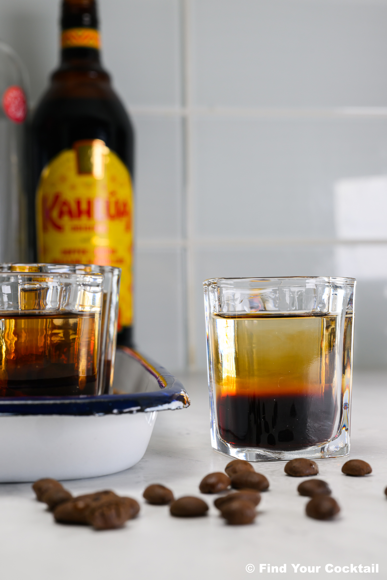 A glass of layered coffee liqueur cocktail sits on a counter with coffee beans, another glass, and a bottle of Kahl&uacute;a in the background.
