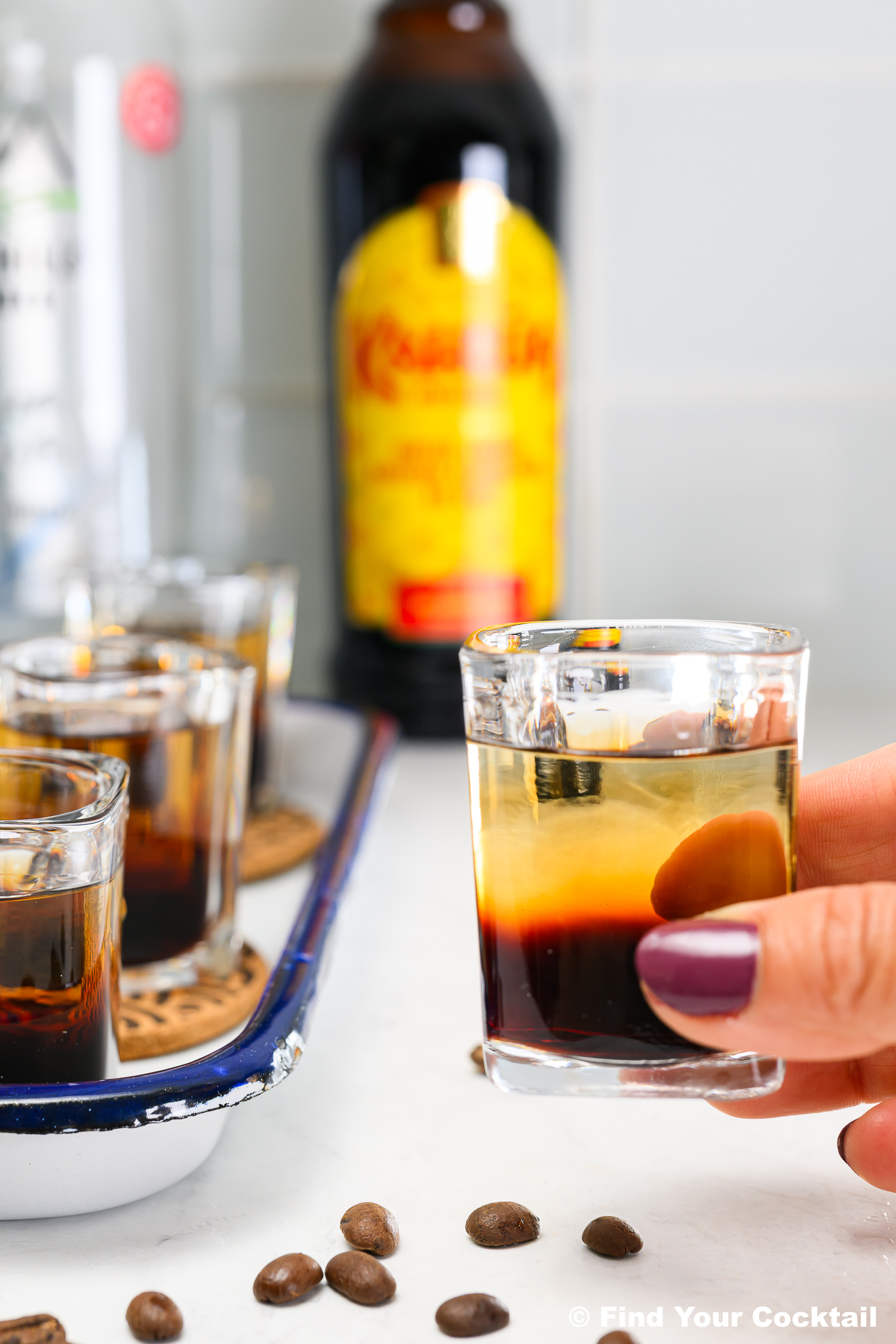 A hand holding a layered shot glass with coffee liqueur and clear spirit; more filled glasses, coffee beans, and bottles are in the background.