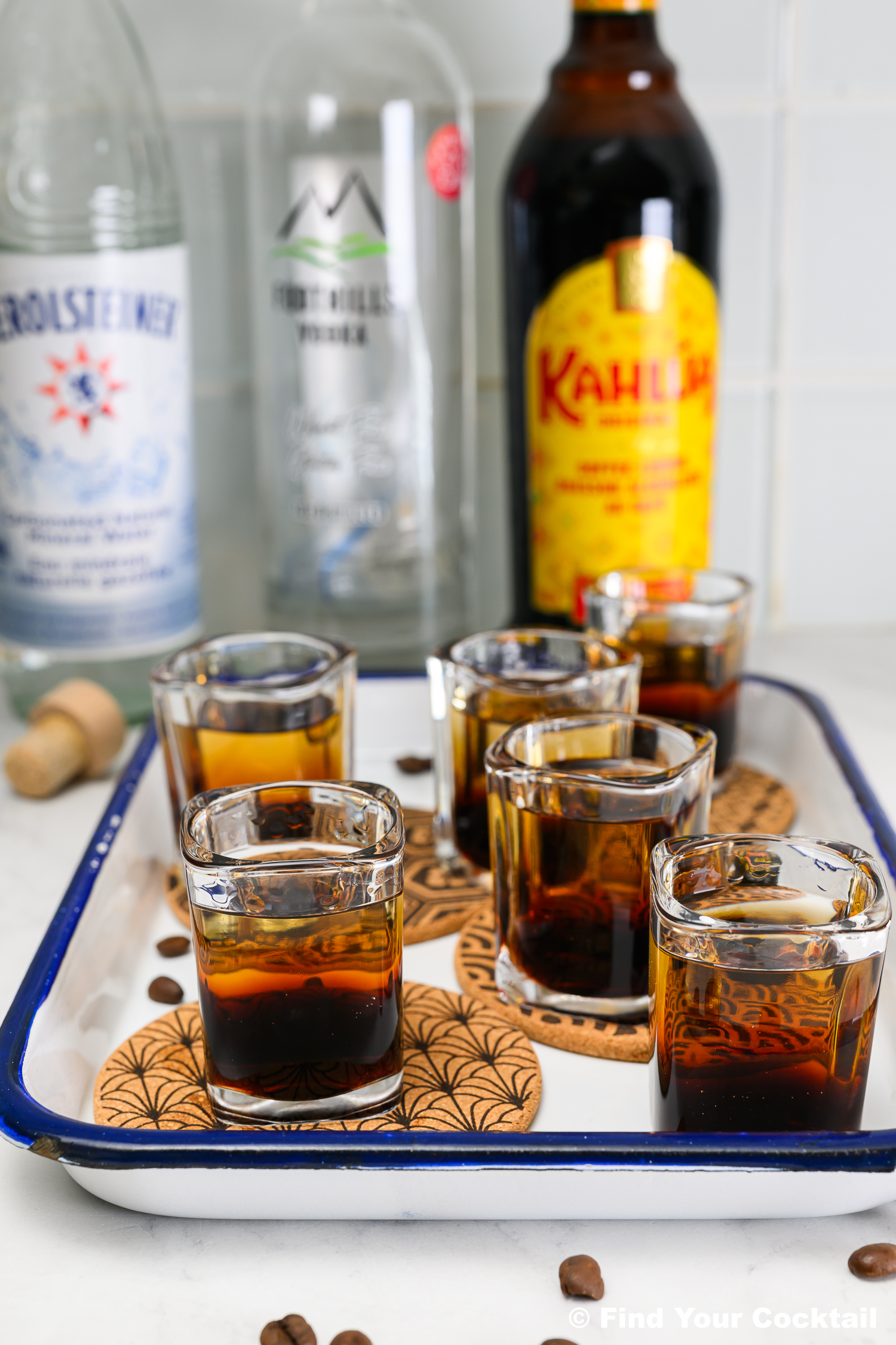 Six shot glasses filled with a dark layered drink are placed on coasters in a tray, with bottles of vodka, coffee liqueur, and water in the background. Coffee beans are scattered nearby.