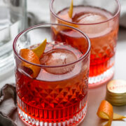 Two glasses of negroni cocktail with large round ice cubes and orange twists on a white surface, next to a bottle and a bottle cap.