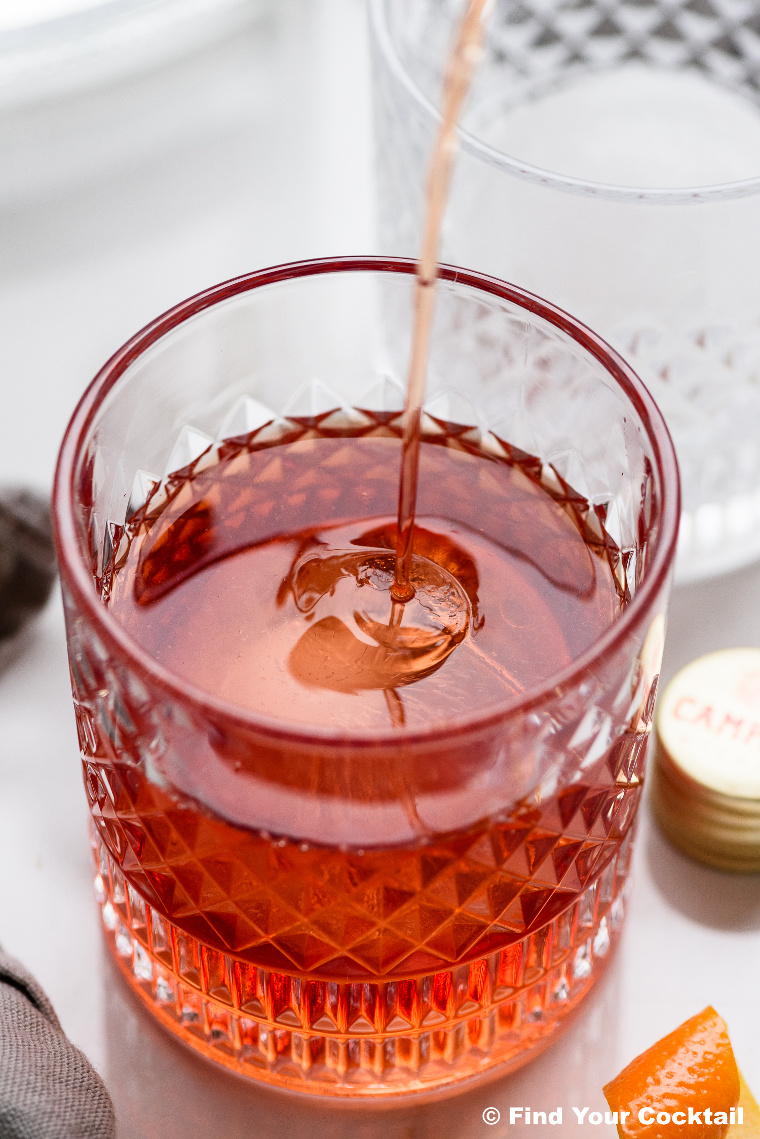 A close-up of a cocktail glass with a clear ice cube as a red liquid is being poured in, with a bottle cap and orange peel nearby.