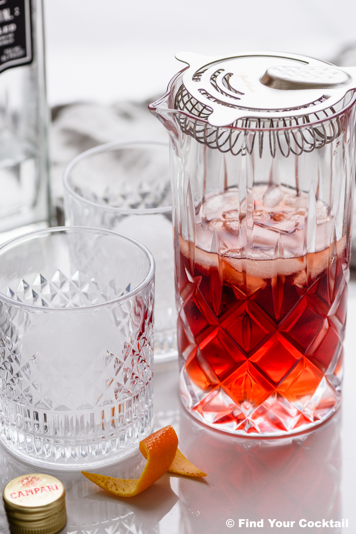 A mixing glass with a strainer holds a red cocktail with ice. Two empty crystal glasses, an orange peel, and a bottle cap are nearby on a white surface.
