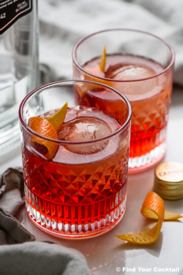 Two glasses of negroni cocktail with large round ice cubes and orange twists on a white surface, next to a bottle and a bottle cap.