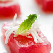 Close-up of a pink, square-shaped Jello shot topped with shredded coconut and a mint leaf, arranged on a white surface.