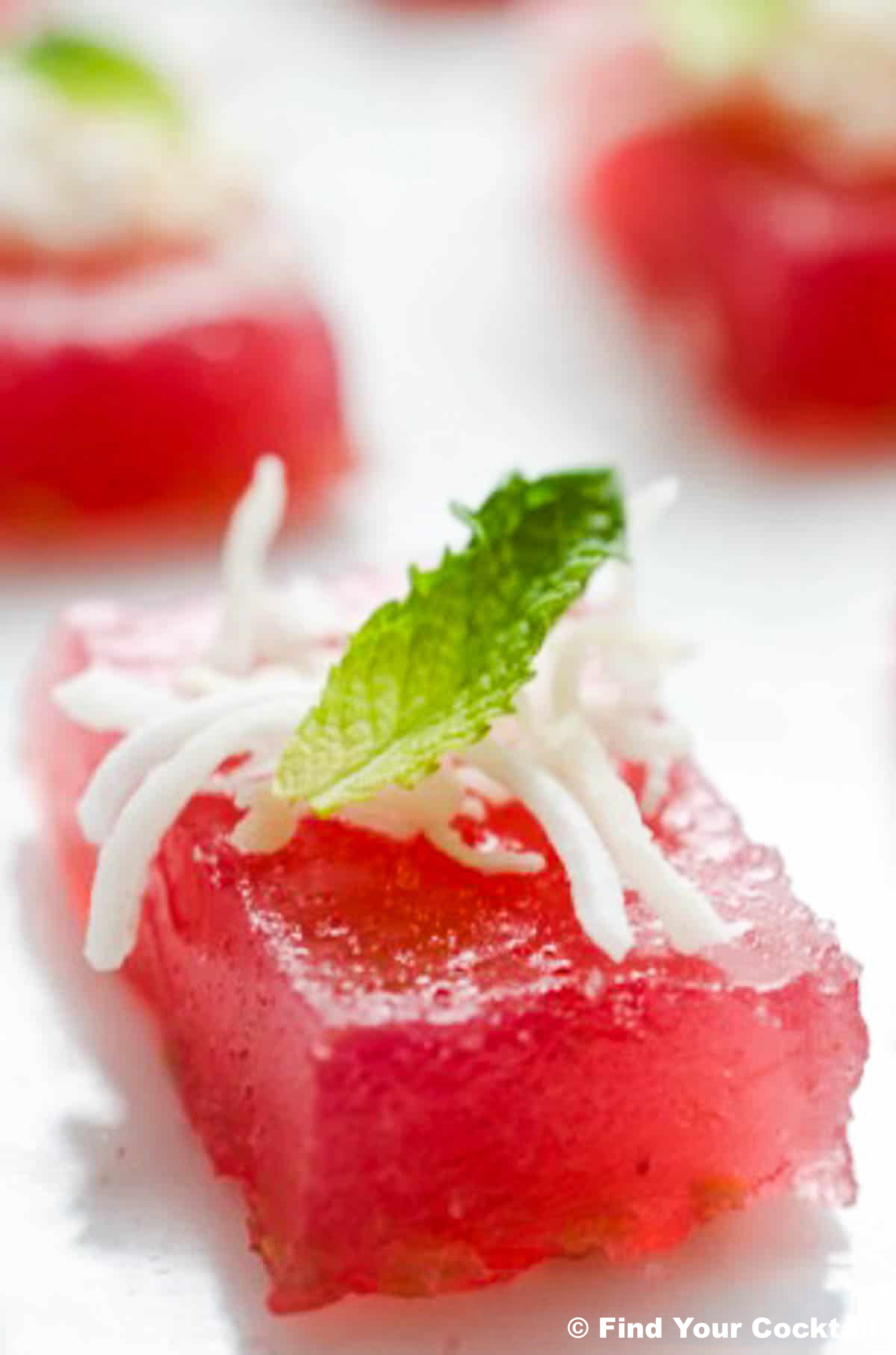 Close-up of a pink, square-shaped Jello shot topped with shredded coconut and a mint leaf, arranged on a white surface.