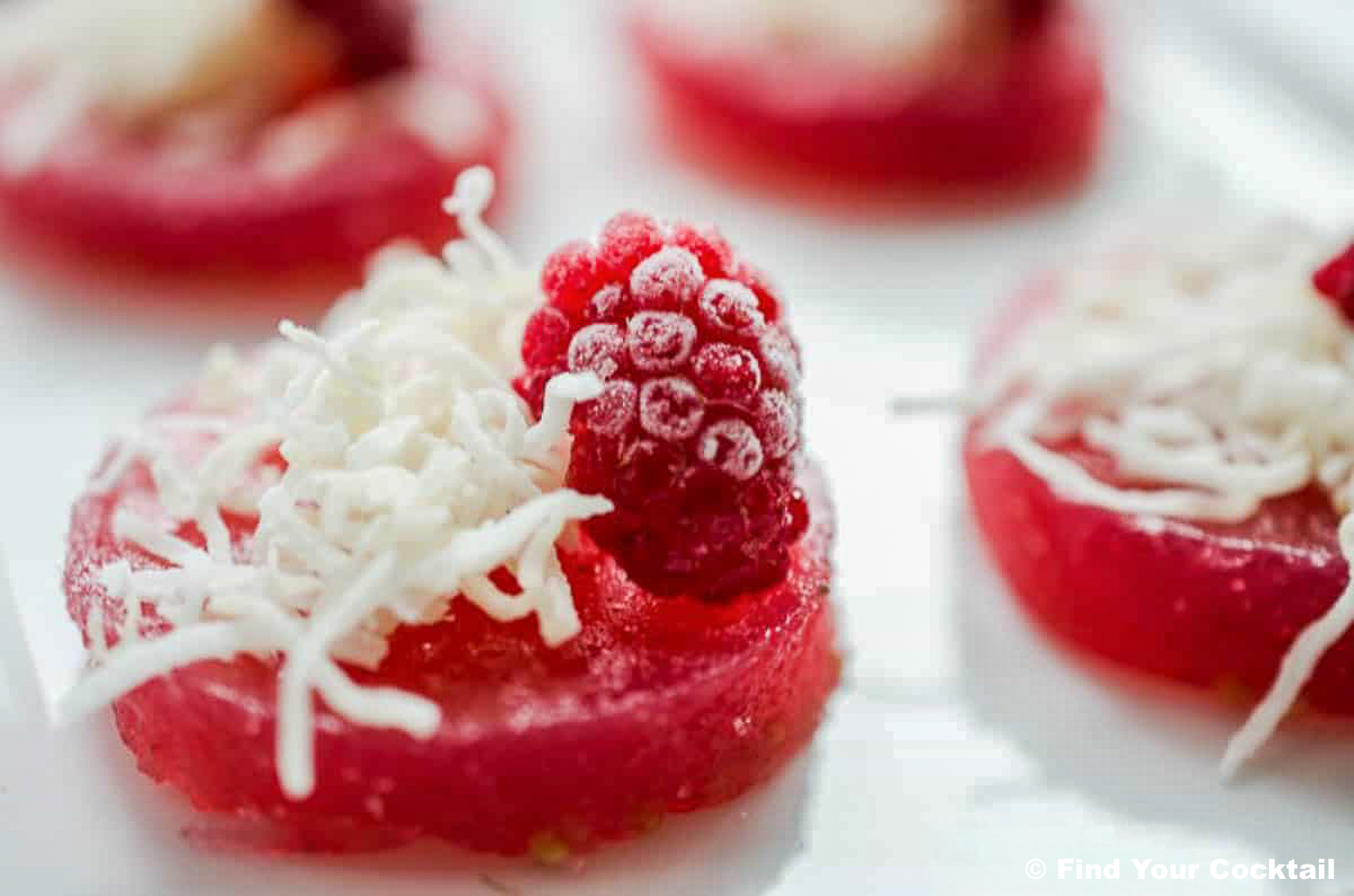 Close-up of a jello shot slice topped with shredded coconut and a raspberry, arranged on a white surface.