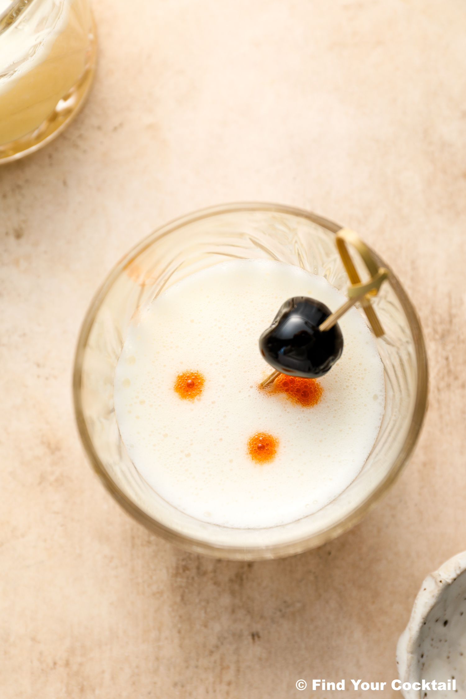 A close-up of a cocktail topped with foam, two drops of bitters, and garnished with a cherry on a cocktail pick.