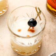 A whiskey sour cocktail in a glass with a frothy top, garnished with a cherry on a cocktail pick and three drops of bitters.