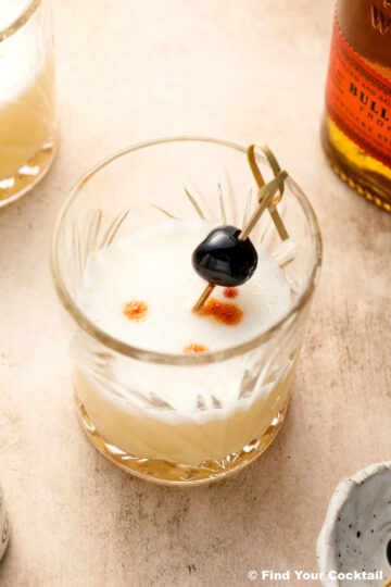 A whiskey sour cocktail in a glass with a frothy top, garnished with a cherry on a cocktail pick and three drops of bitters.