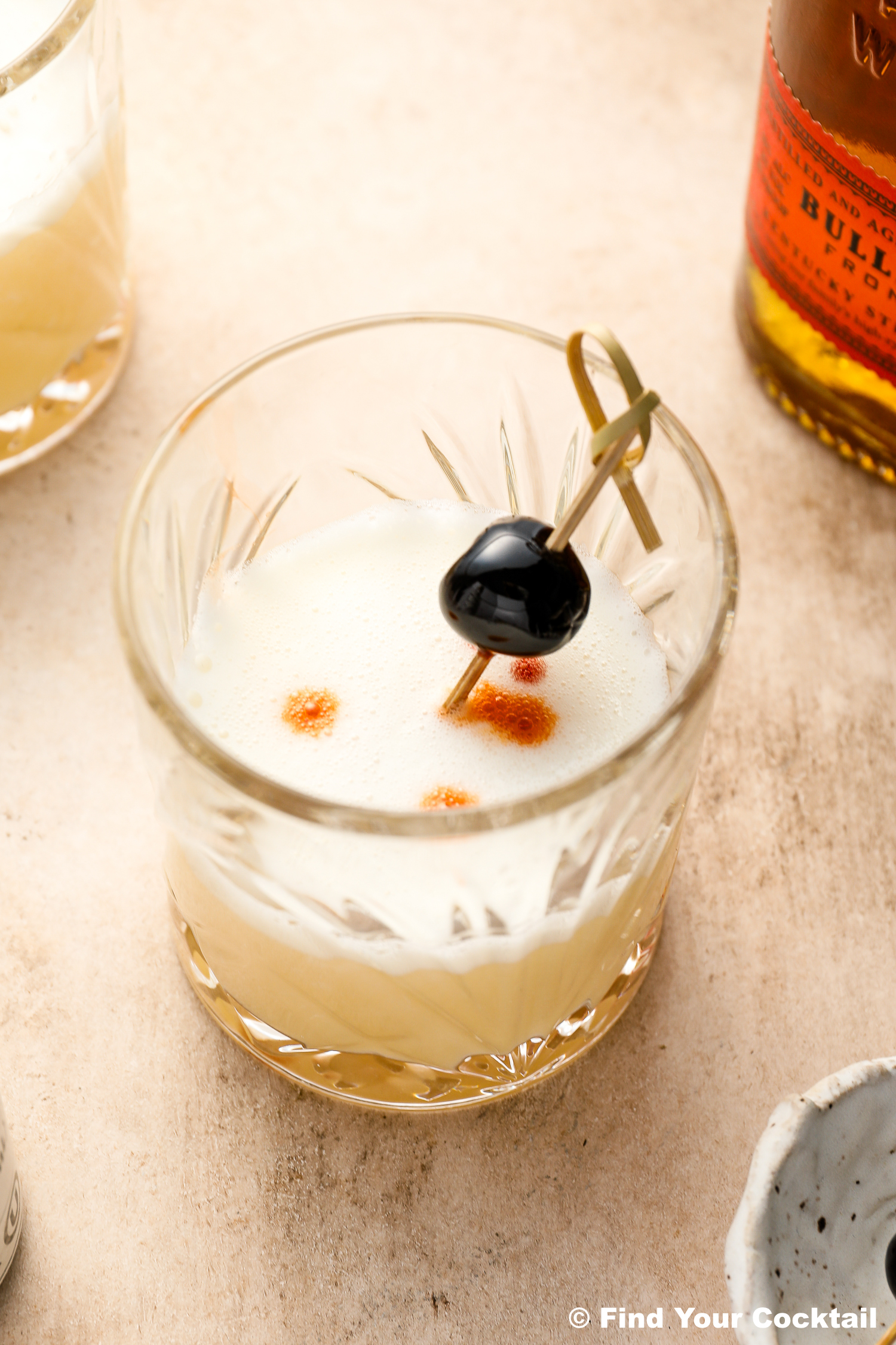 A whiskey sour cocktail in a glass with a frothy top, garnished with a cherry on a cocktail pick and three drops of bitters.