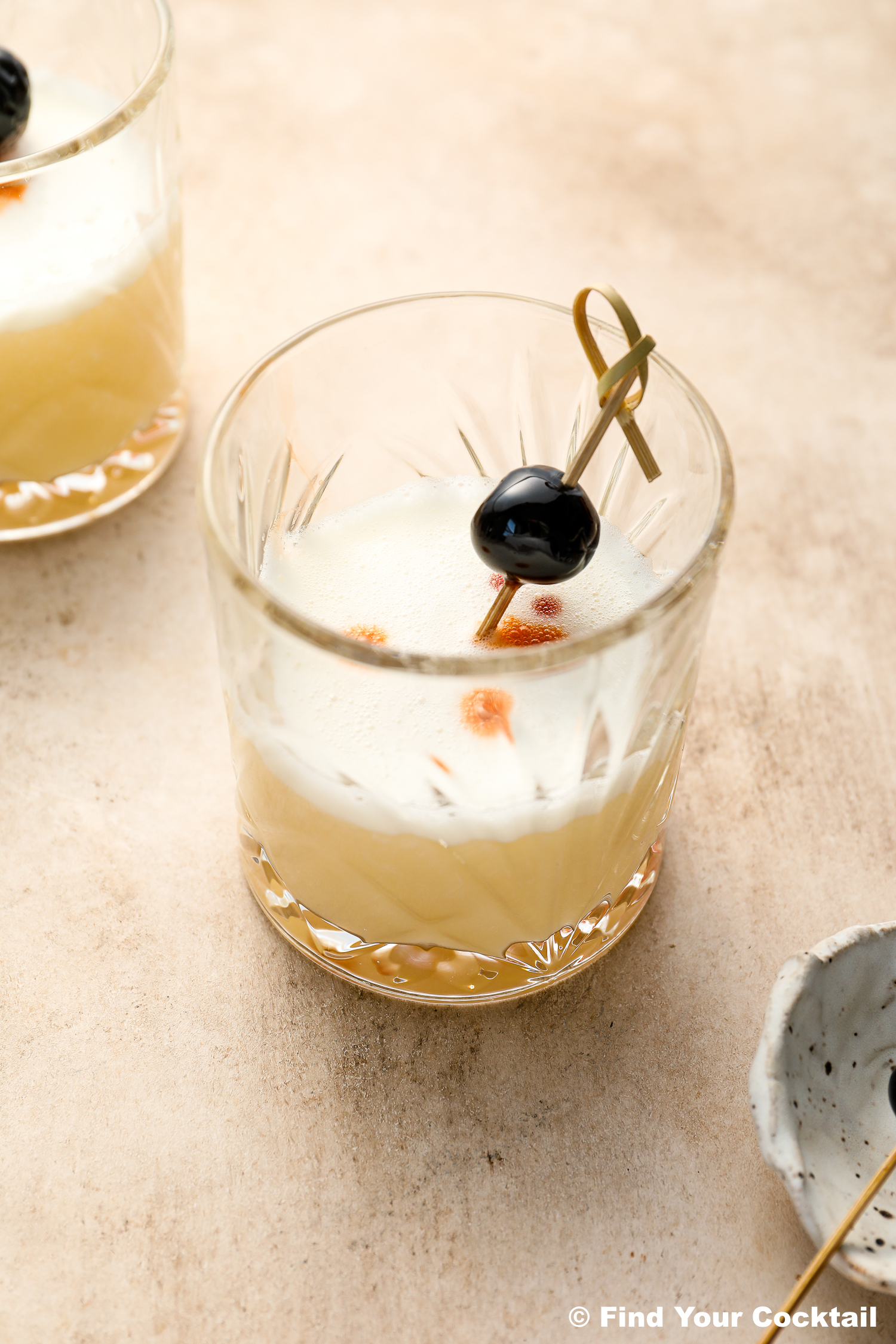 A glass of pale yellow cocktail with a foamy top, garnished with a black cherry on a cocktail pick and a few drops of bitters, sits on a light surface.