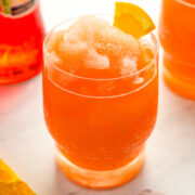 A glass of orange slushy cocktail garnished with an orange wedge sits on a marble surface, with a bottle and orange slice in the background.