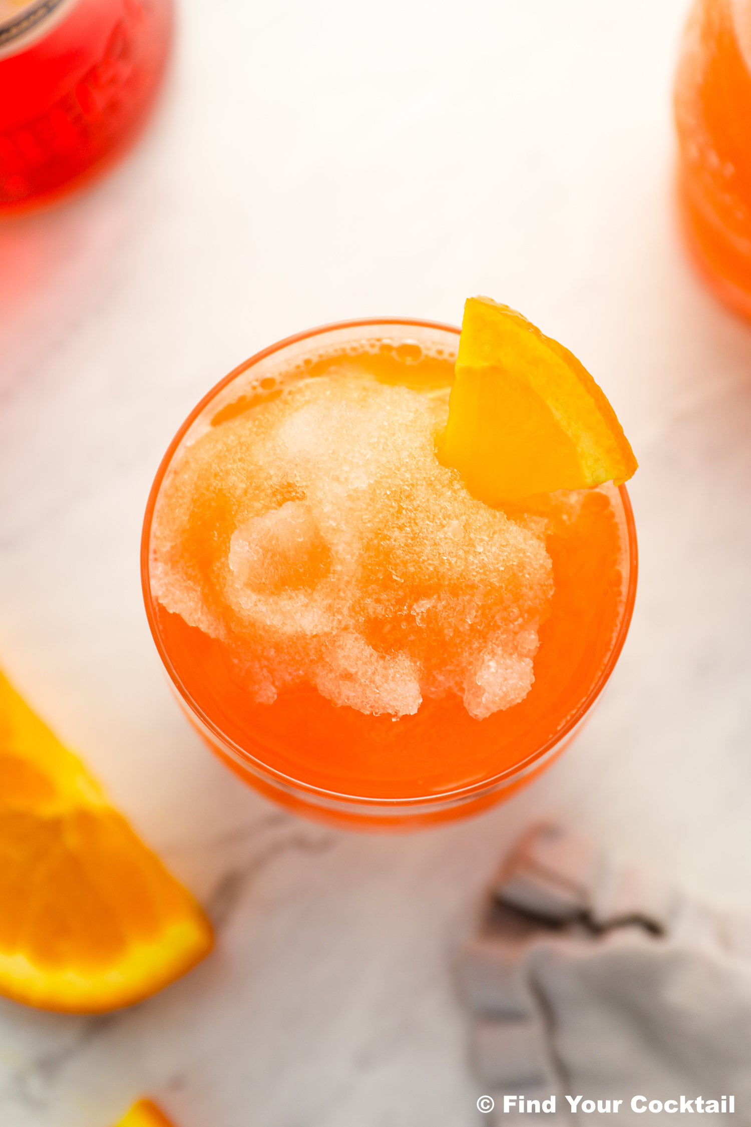 An orange slushy drink in a glass, garnished with an orange wedge, sits on a marble surface with orange slices nearby.