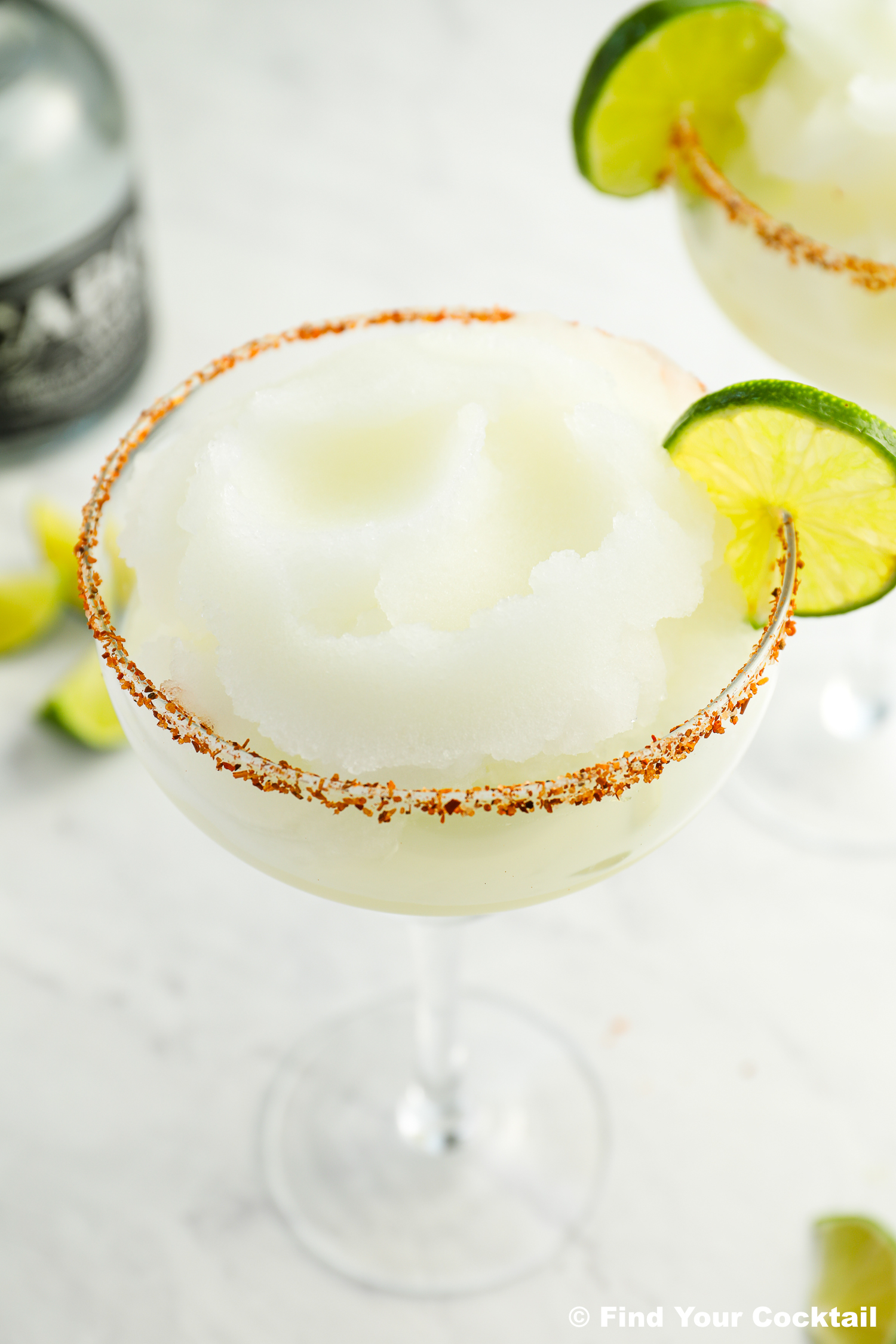 A frozen margarita in a glass with a chili powder rim and lime wheel garnish, set on a light surface with lime wedges and a liquor bottle in the background.