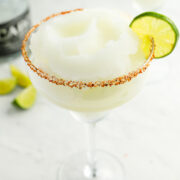 A frozen margarita in a glass with a chili powder rim, garnished with a lime slice. A bottle and lime wedges are in the background.
