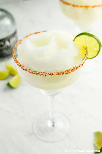 A frozen margarita in a glass with a chili powder rim, garnished with a lime slice. A bottle and lime wedges are in the background.