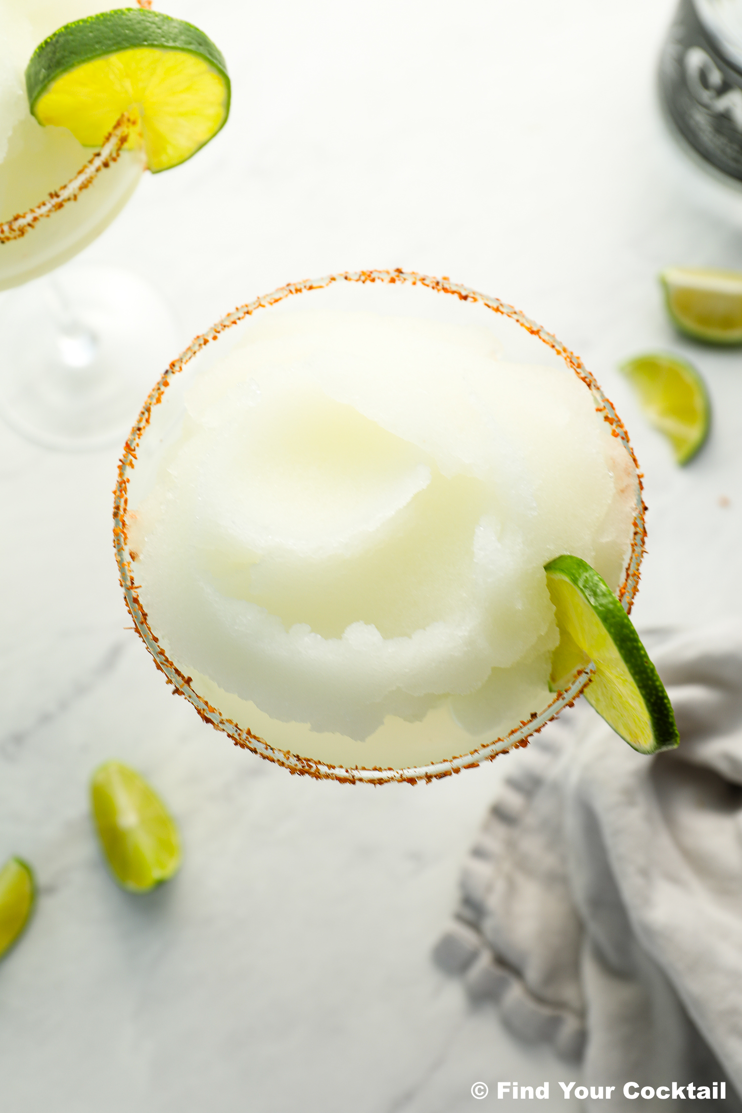 Top view of a frozen margarita in a glass with a chili salt rim, garnished with a lime wedge, surrounded by lime pieces on a light surface.