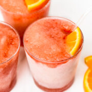 Three glasses of red frozen slush drinks, each garnished with an orange slice, on a white surface.