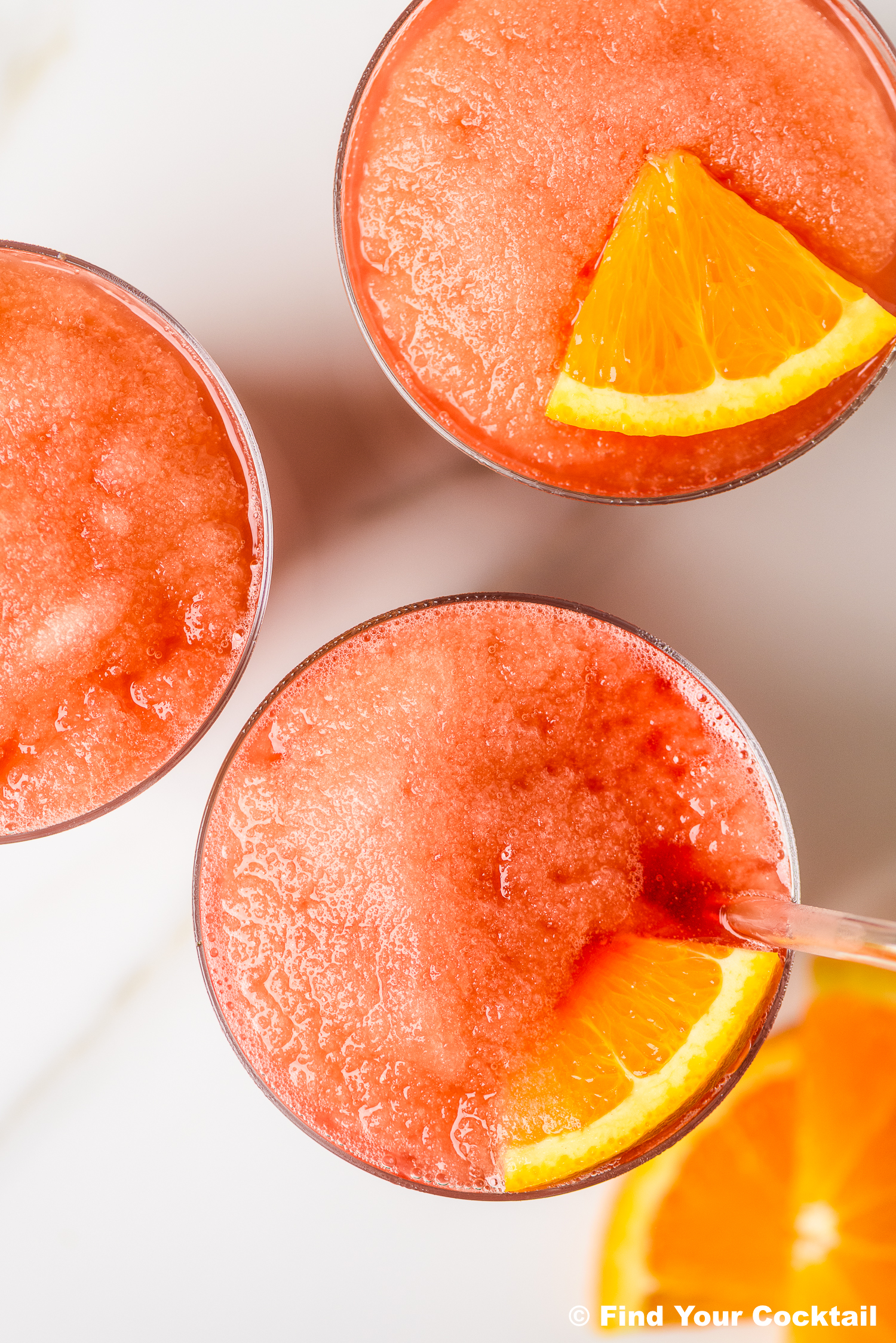 Three glasses of a red slushy drink, each garnished with an orange slice, are shown from above on a white surface.