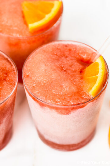 Three glasses of red frozen slush drinks, each garnished with an orange slice, on a white surface.