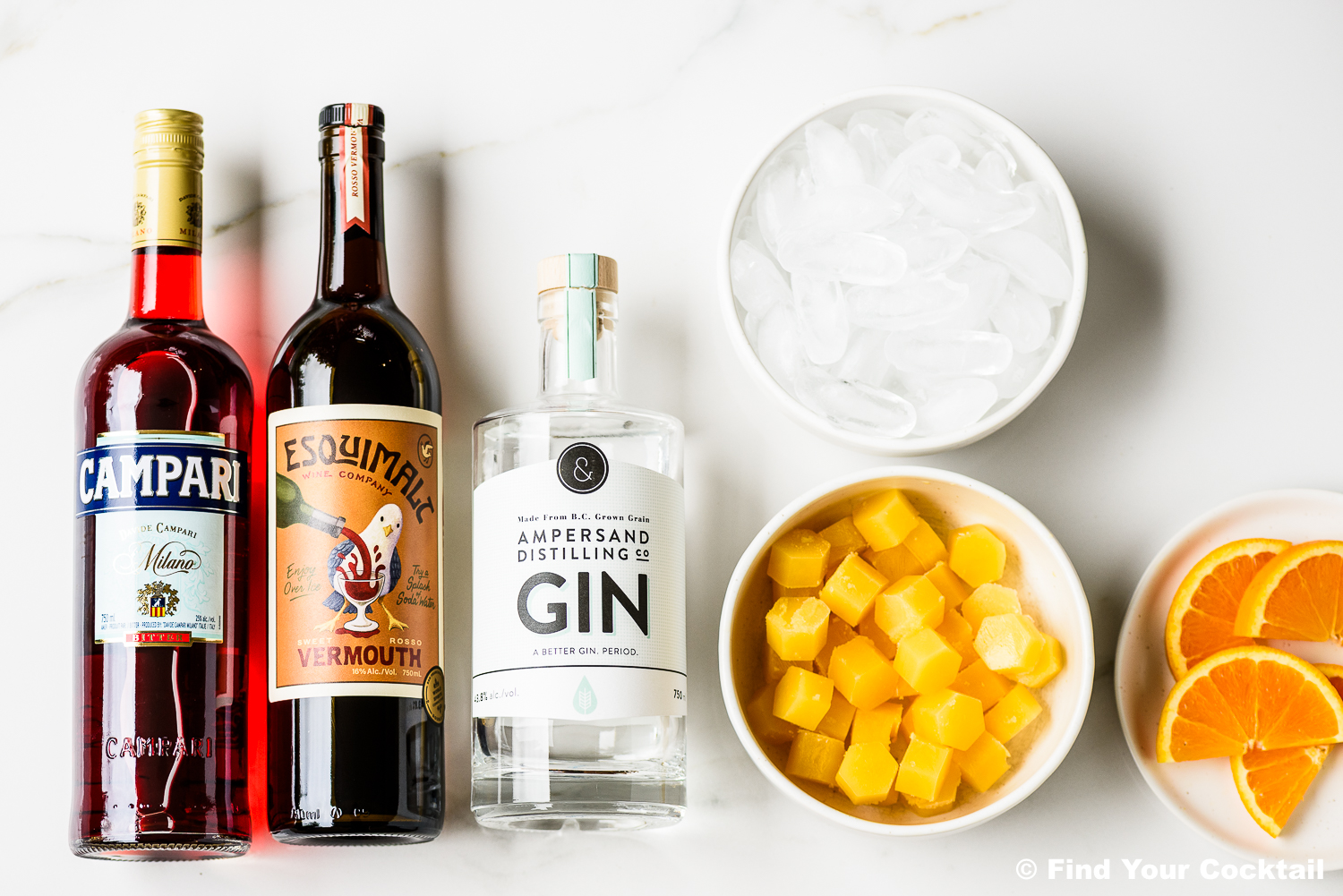 Three bottles (Campari, vermouth, gin) are lined up next to bowls of ice, diced mango, and orange slices on a white surface.