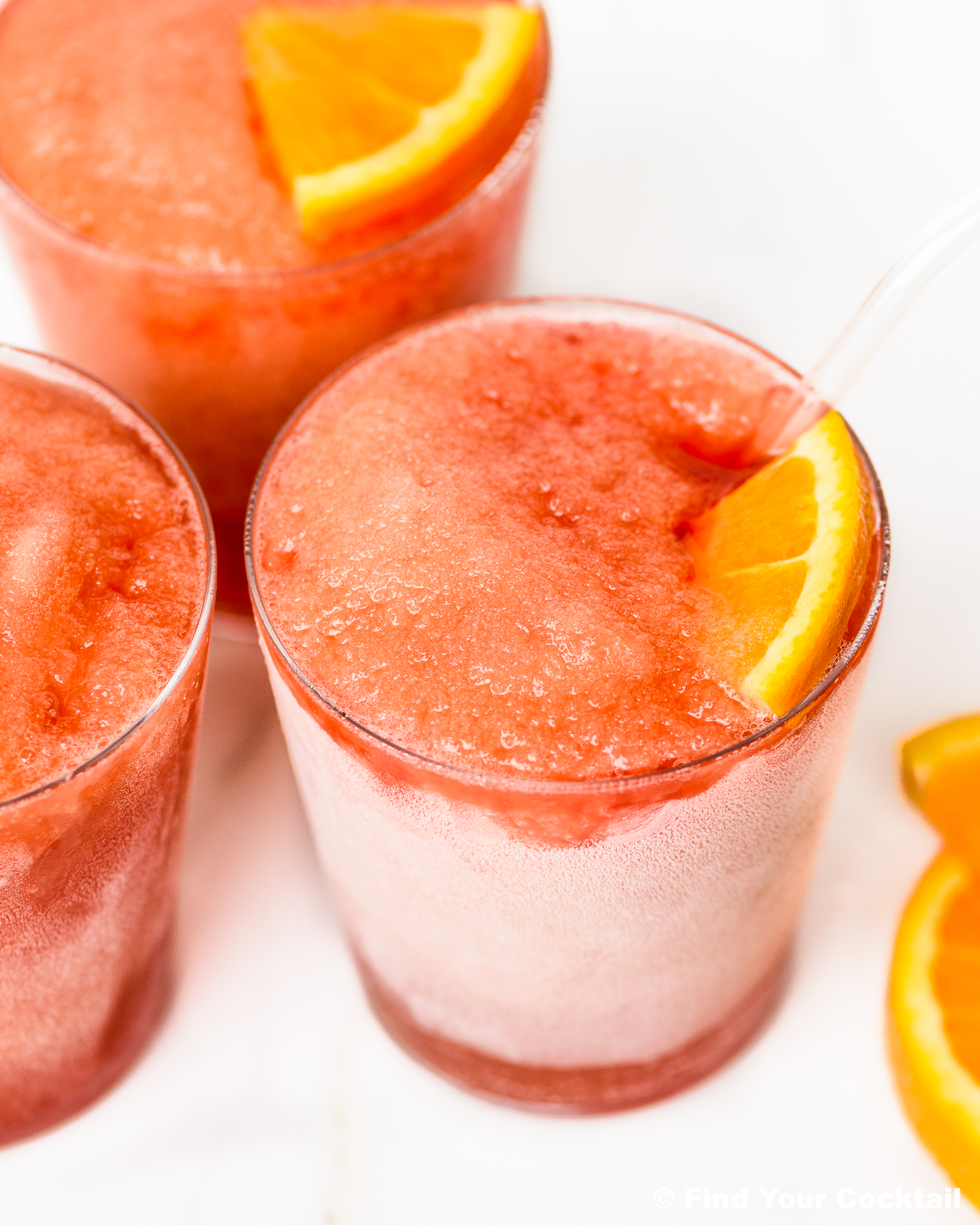 Three glasses of red frozen slush drinks, each garnished with an orange slice, on a white surface.