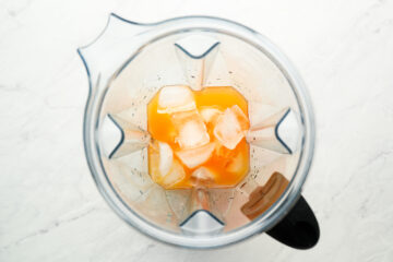 Overhead view of a blender containing orange liquid and several ice cubes on a white surface.