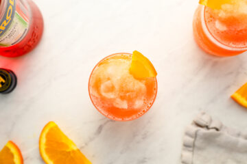 A glass of orange-colored iced cocktail garnished with an orange slice, surrounded by orange wedges and a bottle, on a white marble surface.
