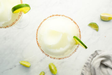Overhead view of a frozen margarita in a glass with a chili-rimmed edge, garnished with a lime wedge; lime slices scattered on a white surface.