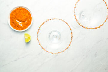 Two empty cocktail glasses with Tajin on the rims, placed beside a bowl of chili powder and a lime wedge on a white marble surface.