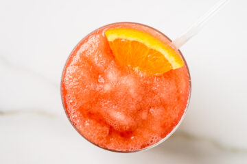 A glass of red slushy drink with crushed ice, garnished with an orange slice and a clear straw, photographed from above on a white surface.