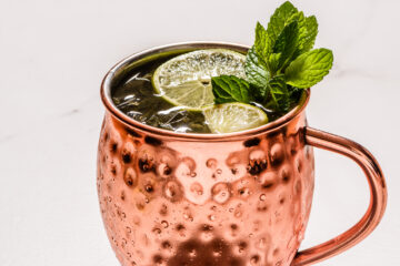 A copper mug filled with a cocktail, garnished with lime slices and fresh mint leaves, against a white background.