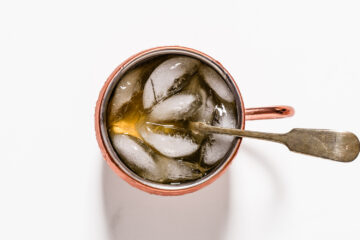 A copper mug filled with a brown beverage, ice cubes, and a metal spoon, photographed from above on a white background.