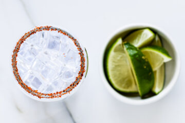 A glass filled with ice and a chili powder rim next to a small bowl containing lime wedges on a white surface.