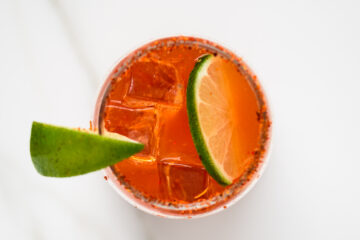 Top-down view of a glass with an orange cocktail, ice cubes, a chili-rimmed edge, and a lime wedge garnish on a white background.