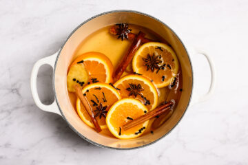 A white pot filled with sliced oranges, cinnamon sticks, star anise, and cloves floating in liquid on a marble surface.