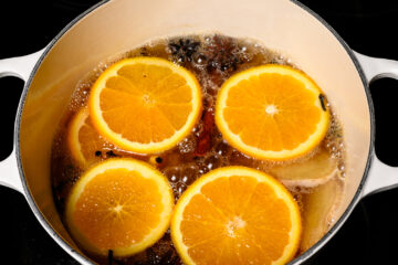 Four orange slices float in a pot of simmering liquid with visible spices, likely being prepared for a beverage or culinary use.
