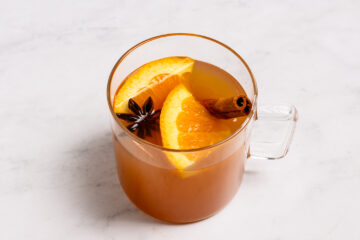 A glass mug filled with a warm orange-colored beverage, garnished with orange slices, a cinnamon stick, and a star anise on a white surface.