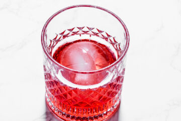 A faceted glass filled with a red beverage and a large spherical ice cube, placed on a white marble surface.
