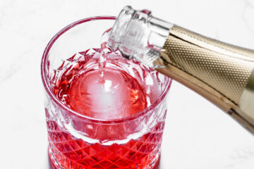 A bottle pours a clear liquid into a glass filled with red liquid and a large round ice cube, on a white surface.
