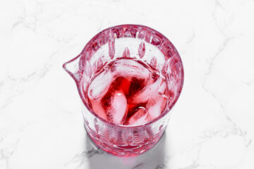 A clear pink glass pitcher filled with red liquid and ice cubes, placed on a white marble surface.