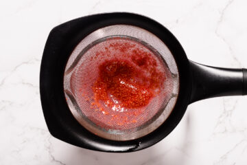 A fine mesh strainer containing mashed strawberries sits over a black bowl on a white marble surface.