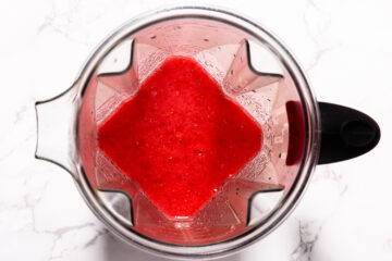Top view of a blender filled with a bright red liquid, possibly a fruit smoothie or juice, on a white marble surface.